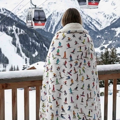 Person seen from behind on the terrace of a mountain chalet, wrapped in a white, soft, and thick blanket adorned with skiers.