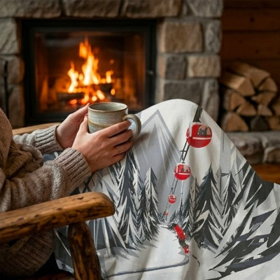 Frau, die in einem Berghaus am Kamin sitzt, eingehüllt in ein Plaid, das eine Skiabfahrt unter Seilbahnen darstellt.